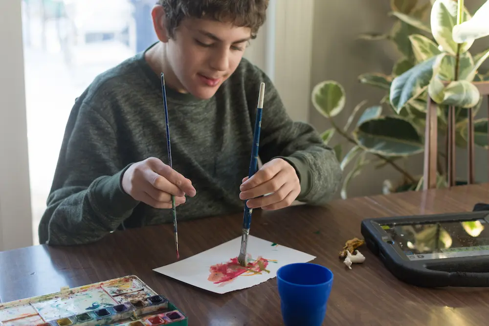 Boy with special needs water color painting with both hands at the same time; AAC communication device sits nearby