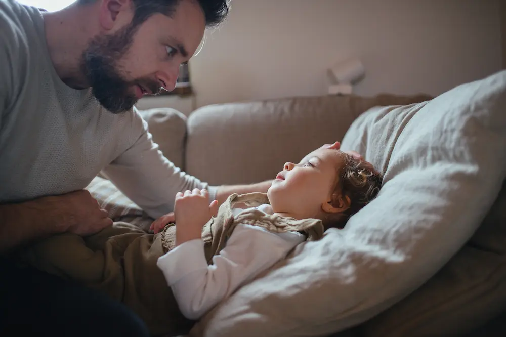 Father taking care of sick girl with fever and cold at home. Daughter lying on sofa, feeling tired.