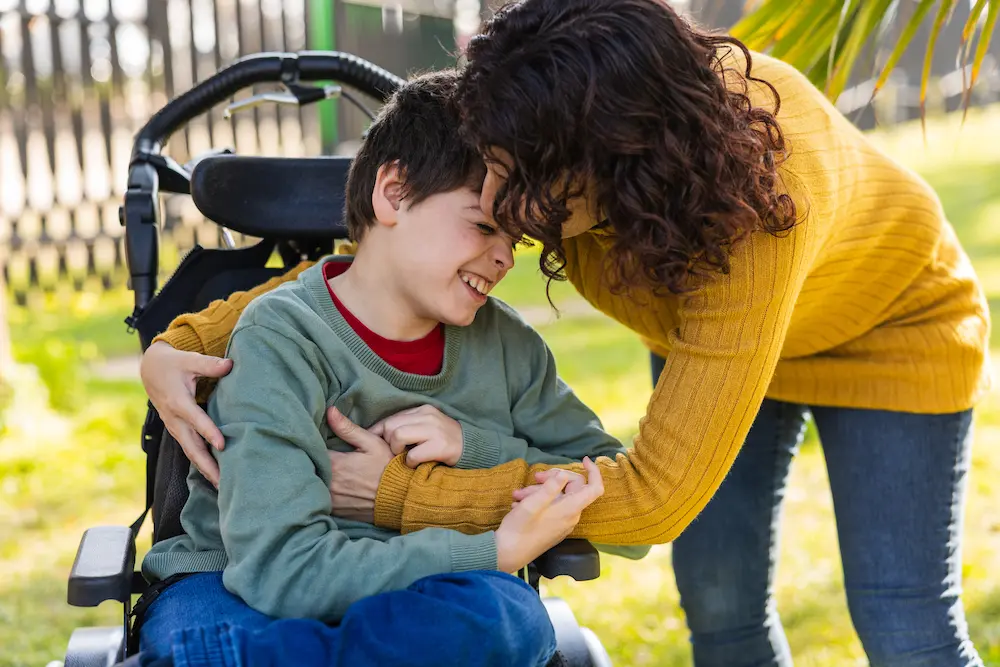 Mother embracing son with disability in wheelchair in park, cerebral palsy awareness