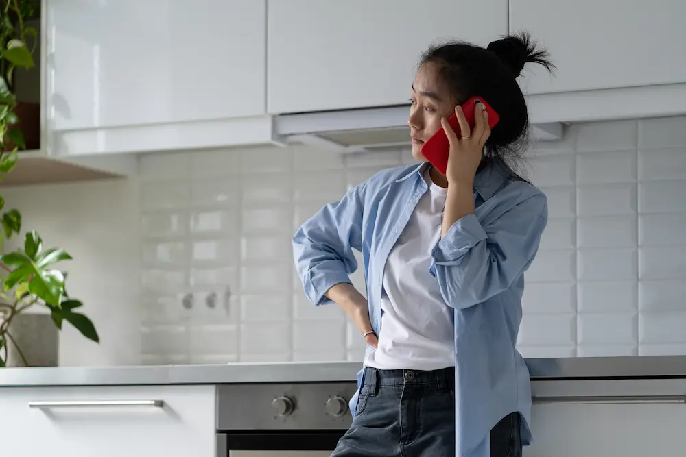 Worried upset Asian woman housewife standing alone in kitchen making unpleasant phone call